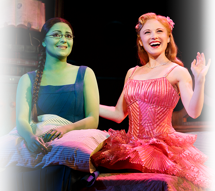 Elphaba and Glinda Sitting on a Bed Smiling